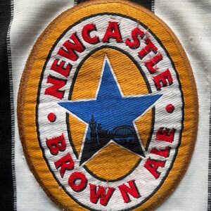 May include: A circular patch with the words "Newcastle Brown Ale" in red lettering. The patch features a blue star with a silhouette of a bridge and buildings. The patch is on a black and white striped fabric.