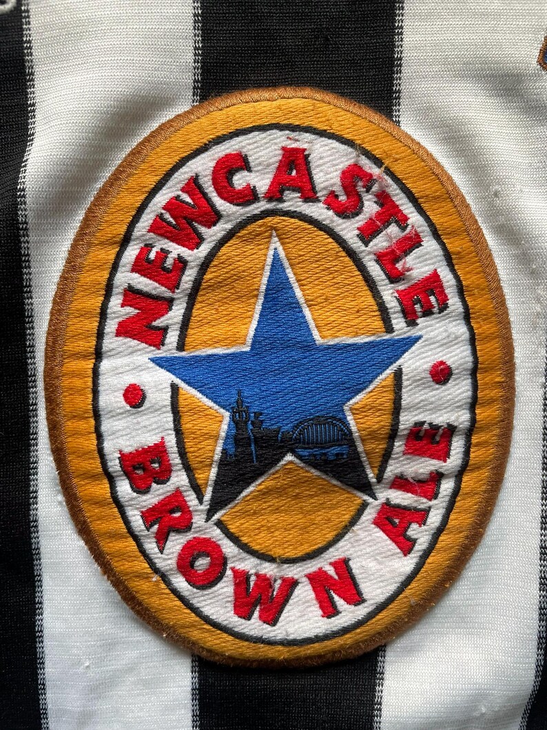 May include: A circular patch with the words "Newcastle Brown Ale" in red lettering. The patch features a blue star with a silhouette of a bridge and buildings. The patch is on a black and white striped fabric.