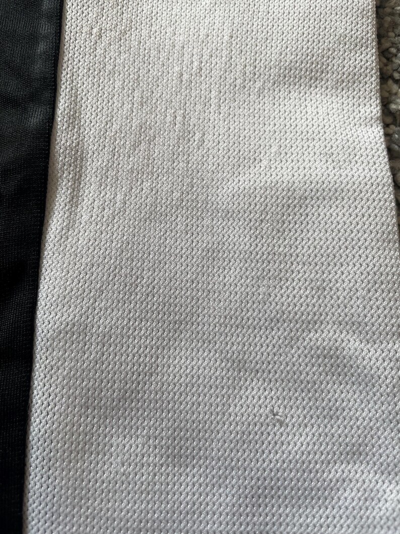 May include: Close-up of a white fabric with a textured, woven pattern. The fabric has a black stripe running down the left side.