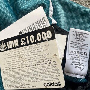 May include: Close-up of a teal and black athletic jersey with white accents. Attached tags include a contest entry card with "WIN &pound;10,000" and an Adidas label. The jersey has a textured pattern.