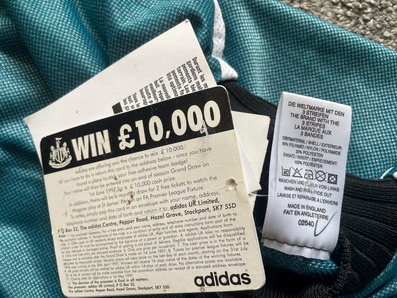 May include: Close-up of a teal and black athletic jersey with white accents. Attached tags include a contest entry card with "WIN &pound;10,000" and an Adidas label. The jersey has a textured pattern.