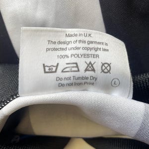 May include: A white tag with black text, including "Made in U.K." and care instructions. The tag is attached to a black and white striped fabric, likely part of a garment. The fabric is 100% polyester.