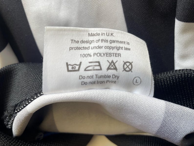 May include: A white tag with black text, including "Made in U.K." and care instructions. The tag is attached to a black and white striped fabric, likely part of a garment. The fabric is 100% polyester.