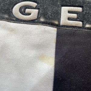 May include: Close-up of a black and white fabric with the letters "GE" in white, raised lettering. The letters are on a dark gray, textured background. The fabric has a white, textured weave on the left side and a black, smooth weave on the right side.