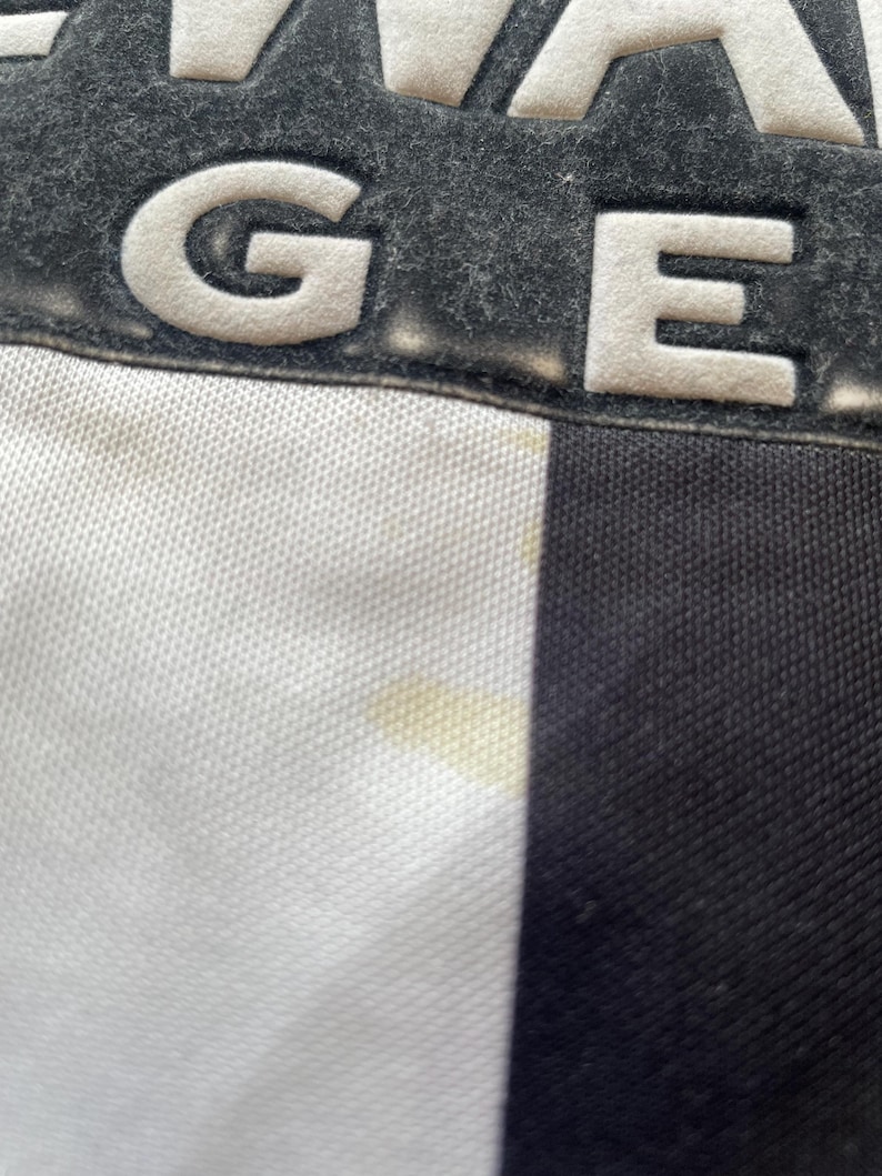 May include: Close-up of a black and white fabric with the letters "GE" in white, raised lettering. The letters are on a dark gray, textured background. The fabric has a white, textured weave on the left side and a black, smooth weave on the right side.
