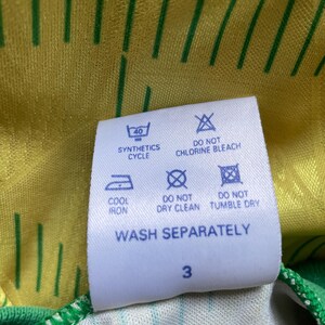 May include: White care label with blue text and symbols. The label includes instructions for washing, drying, and ironing a garment. The text reads "Synthetics Cycle", "Do Not Chlorine Bleach", "Cool Iron", "Do Not Dry Clean", "Do Not Tumble Dry", and "Wash Separately". The number "3" is printed at the bottom of the label.