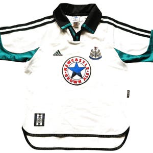 May include: White soccer jersey with black and green accents. The jersey has the Adidas logo, the Newcastle United crest, and a circular logo that says "Newcastle Brown Ale" with a star in the center.