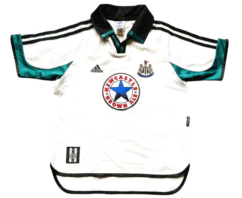 May include: White soccer jersey with black and green accents. The jersey has the Adidas logo, the Newcastle United crest, and a circular logo that says "Newcastle Brown Ale" with a star in the center.