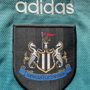 May include: A teal soccer jersey with the Adidas logo embroidered in white. The Newcastle United team crest is prominently displayed, featuring two seahorses and the text "Newcastle United" in blue.