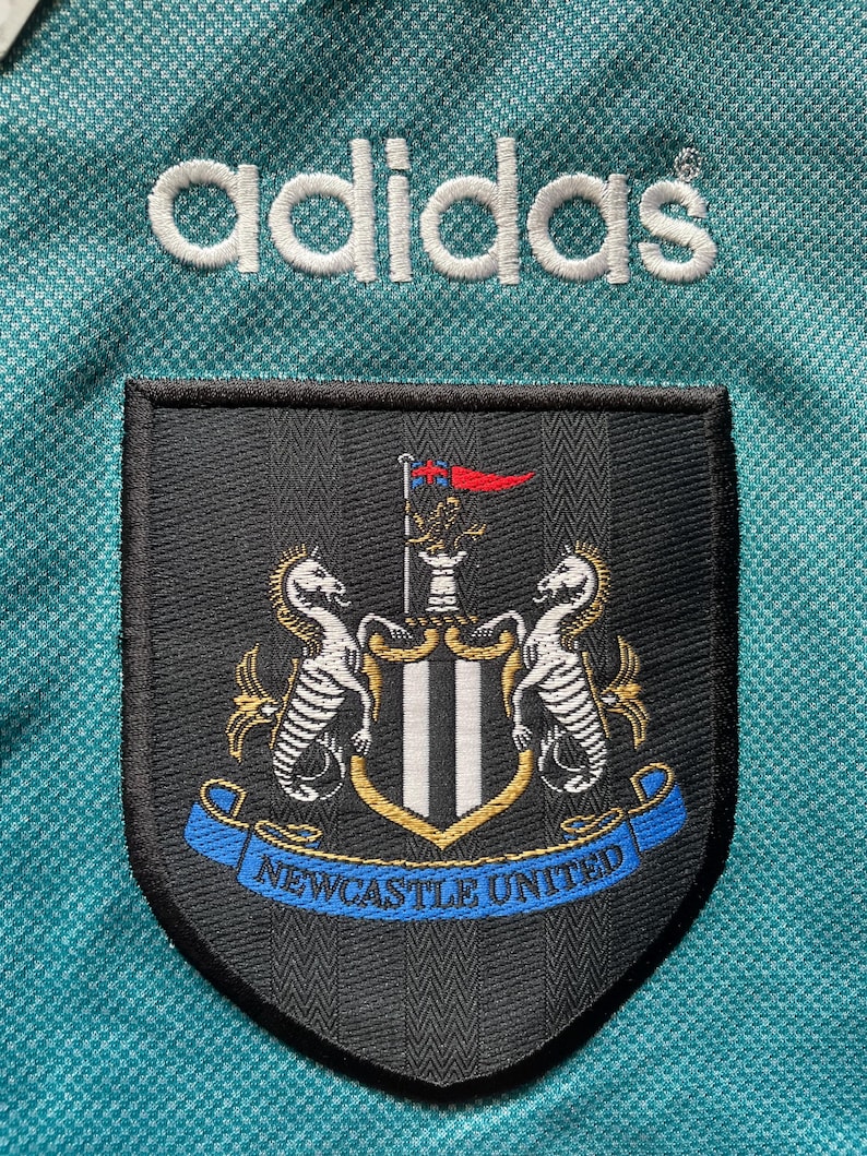 May include: A teal soccer jersey with the Adidas logo embroidered in white. The Newcastle United team crest is prominently displayed, featuring two seahorses and the text "Newcastle United" in blue.