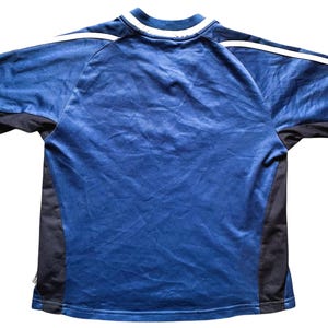 Op de afbeelding: Een blauw sportshirt met zwarte zijpanelen en mouwen. Witte strepen lopen langs de schouders. De kraag is wit met een blauwe bies. Het shirt is gemaakt van lichtgewicht, ademend materiaal.