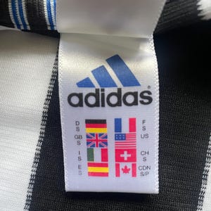 May include: Close-up of a white tag with the Adidas logo in blue. The tag displays the brand name and various country flags, including the United Kingdom, France, and the United States. The tag is attached to a black and white striped fabric.
