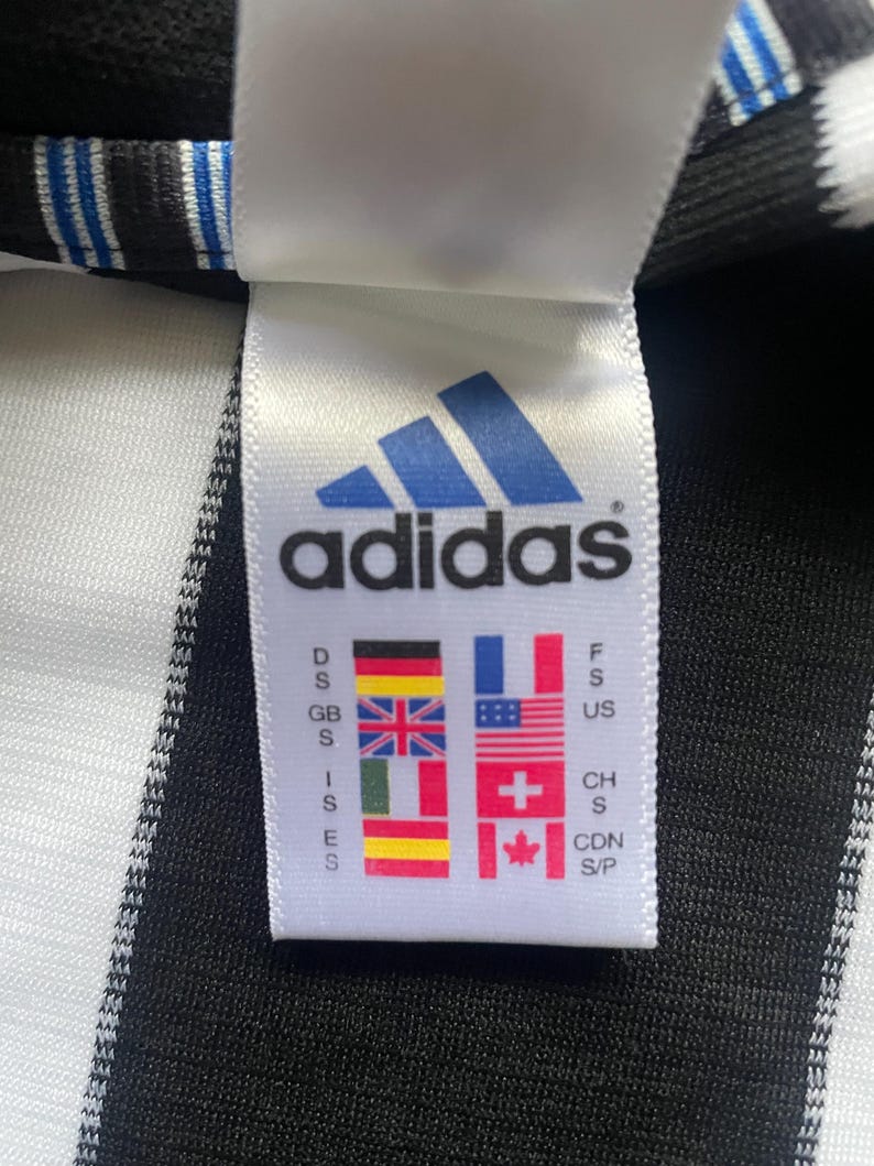 May include: Close-up of a white tag with the Adidas logo in blue. The tag displays the brand name and various country flags, including the United Kingdom, France, and the United States. The tag is attached to a black and white striped fabric.