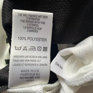 Puede incluir: Primer plano de una etiqueta de prenda blanca con texto negro. La etiqueta dice "DIE WELTMARKE MIT DEN 3 STREIFEN" y "THE BRAND WITH THE 3 STRIPES". La etiqueta tambi&eacute;n indica "100% POLYESTER" y "MADE IN U.K."