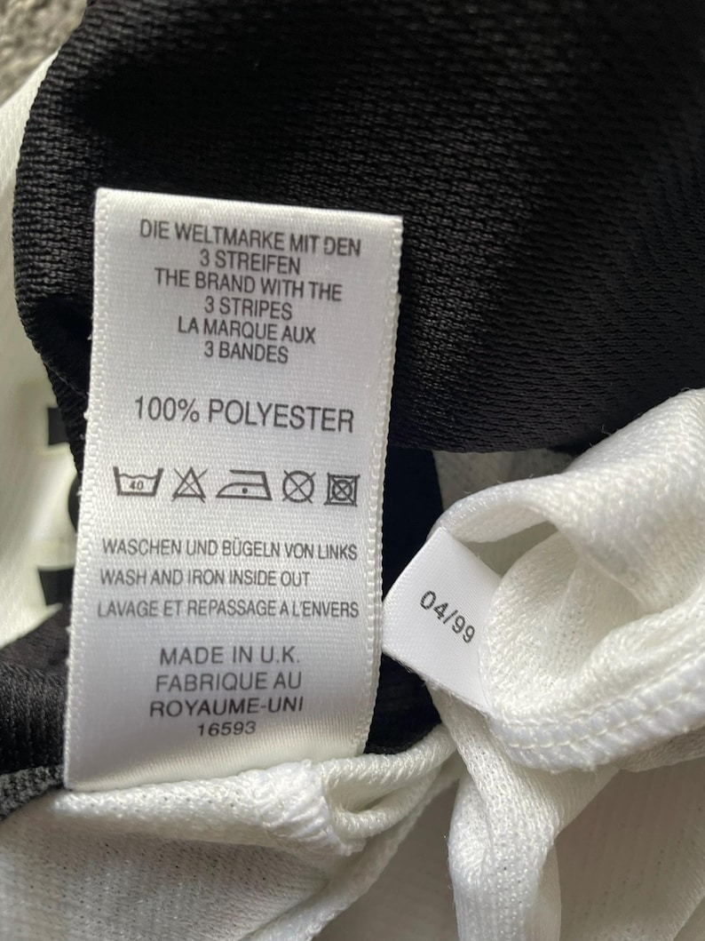 Puede incluir: Primer plano de una etiqueta de prenda blanca con texto negro. La etiqueta dice "DIE WELTMARKE MIT DEN 3 STREIFEN" y "THE BRAND WITH THE 3 STRIPES". La etiqueta tambi&eacute;n indica "100% POLYESTER" y "MADE IN U.K."