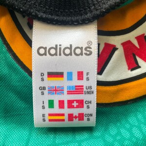 May include: Close-up of an Adidas tag on a green and yellow jersey. The tag displays the Adidas logo and various country flags, including the US, UK, and Canada, along with size indicators.
