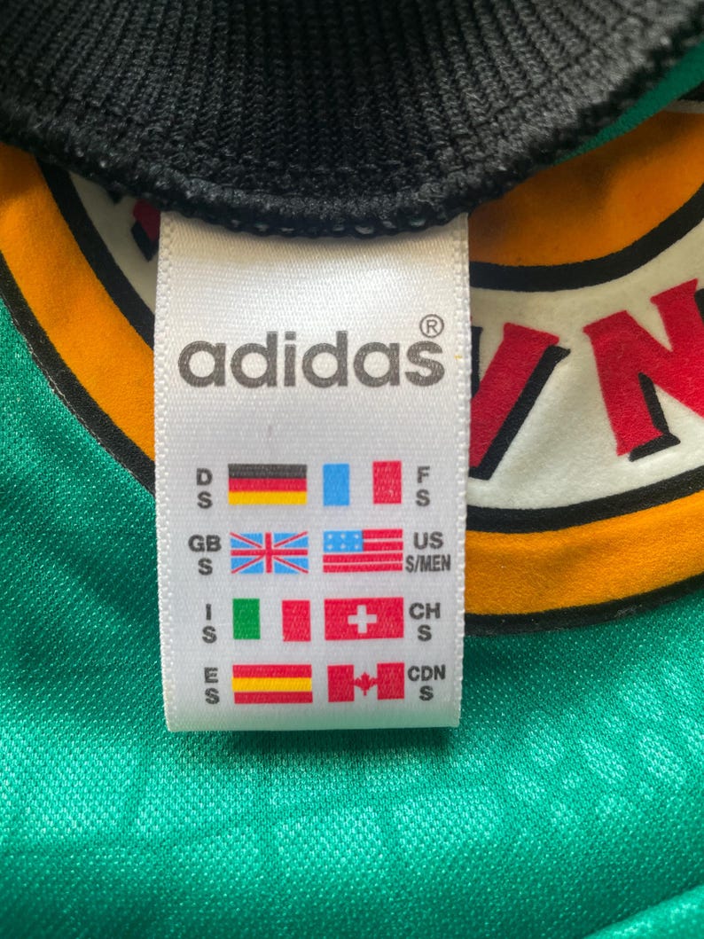 May include: Close-up of an Adidas tag on a green and yellow jersey. The tag displays the Adidas logo and various country flags, including the US, UK, and Canada, along with size indicators.