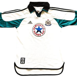 May include: White football shirt with black and teal detailing. Includes a Newcastle Brown Ale logo and Adidas branding. The shirt has a black collar and trim, with a v-neck design.