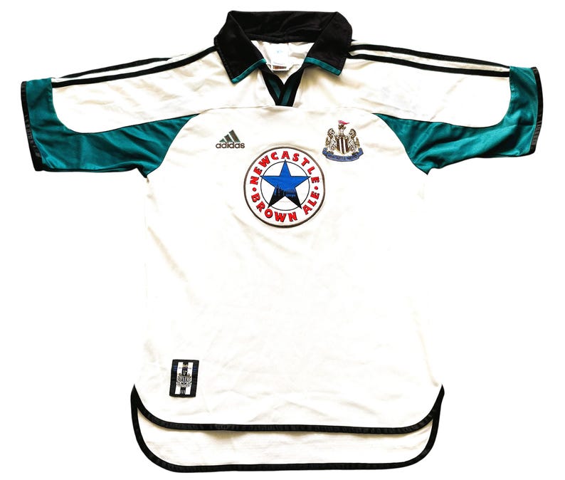 May include: White football shirt with black and teal detailing. Includes a Newcastle Brown Ale logo and Adidas branding. The shirt has a black collar and trim, with a v-neck design.