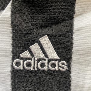 May include: Close-up of a black and white striped fabric with the Adidas logo embroidered in white. The logo features three white stripes above the word "adidas". The fabric has a textured appearance.
