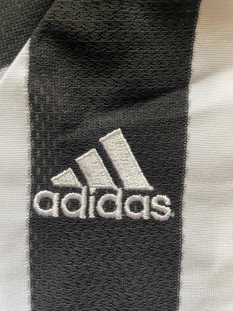 May include: Close-up of a black and white striped fabric with the Adidas logo embroidered in white. The logo features three white stripes above the word "adidas". The fabric has a textured appearance.