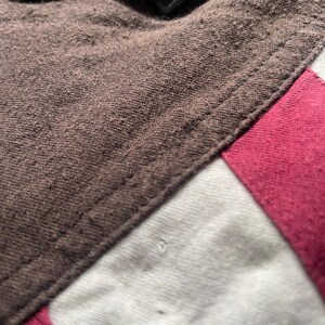 Puede incluir: Primer plano de una tela marr&oacute;n con una costura, que pasa a tela blanca y rosa. Un parche negro con detalles azules y dorados es visible en la parte superior. Las texturas y los colores sugieren una prenda o un producto textil.