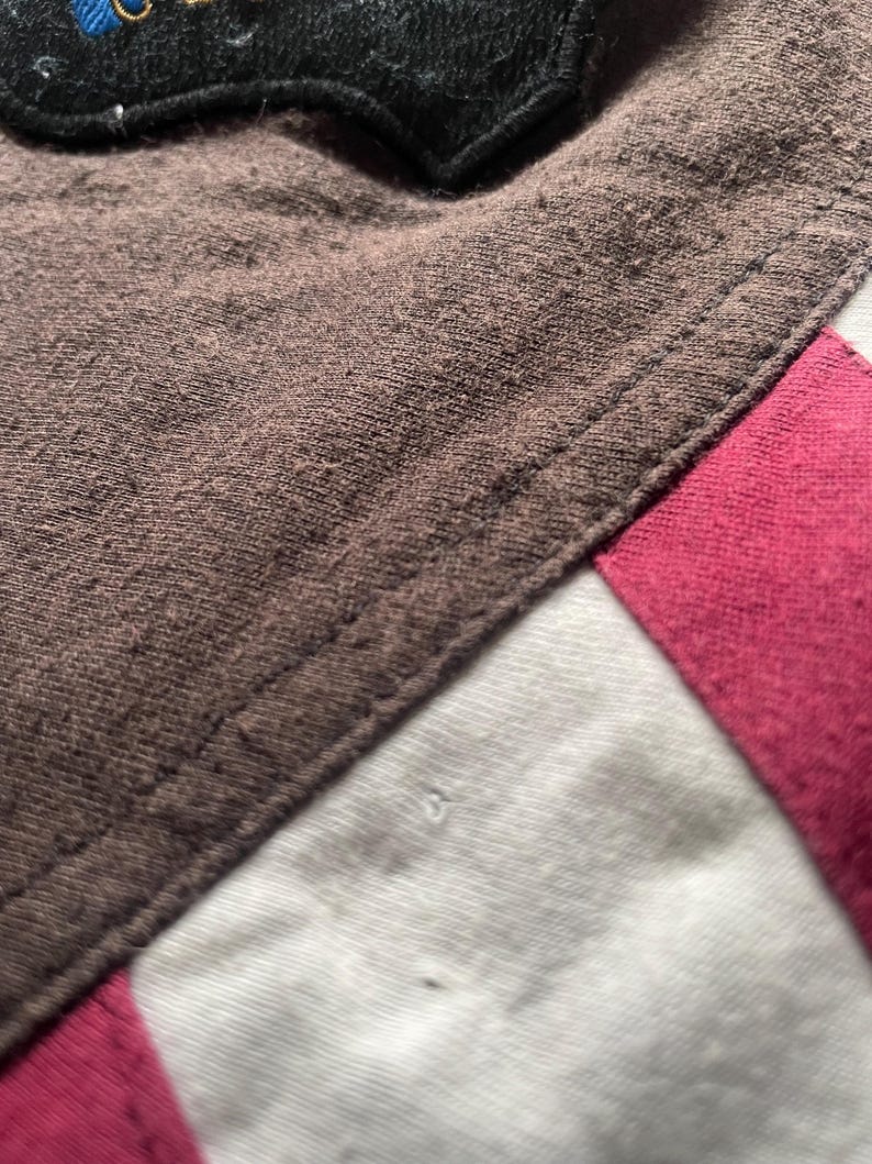 Puede incluir: Primer plano de una tela marr&oacute;n con una costura, que pasa a tela blanca y rosa. Un parche negro con detalles azules y dorados es visible en la parte superior. Las texturas y los colores sugieren una prenda o un producto textil.