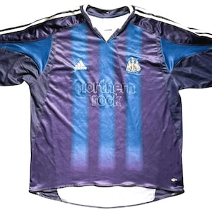 May include: A Newcastle United soccer jersey in navy blue, purple, and light blue. The jersey features the Adidas logo, the club crest, and the text "northern rock". White stripes run down the sleeves.
