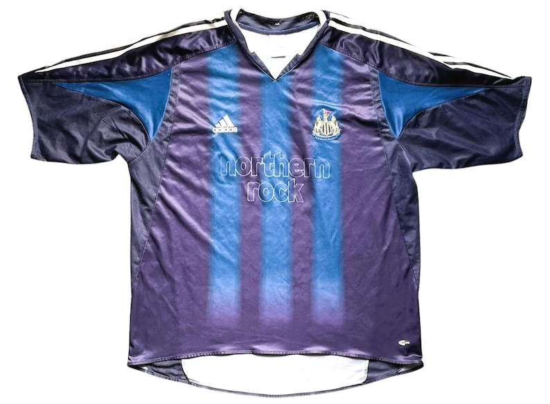 May include: A Newcastle United soccer jersey in navy blue, purple, and light blue. The jersey features the Adidas logo, the club crest, and the text "northern rock". White stripes run down the sleeves.