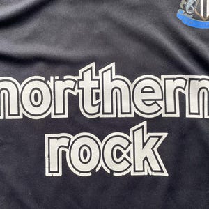 Pu&ograve; includere: Maglia da calcio nera con la scritta "northern rock" a grandi lettere bianche. Lo stemma del Newcastle United &egrave; visibile in alto a destra. Il tessuto sembra leggero e sportivo.