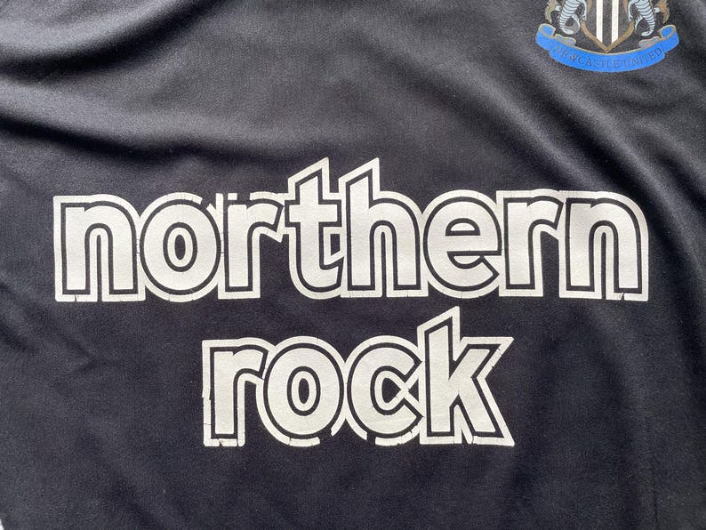 Pu&ograve; includere: Maglia da calcio nera con la scritta "northern rock" a grandi lettere bianche. Lo stemma del Newcastle United &egrave; visibile in alto a destra. Il tessuto sembra leggero e sportivo.