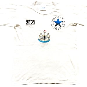 May include: White t-shirt featuring a Newcastle Breweries logo, a blue star, and the Newcastle United Football Club crest. The shirt also has an Asics logo.