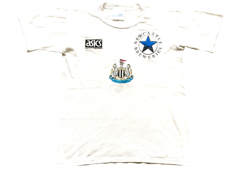 May include: White t-shirt featuring a Newcastle Breweries logo, a blue star, and the Newcastle United Football Club crest. The shirt also has an Asics logo.