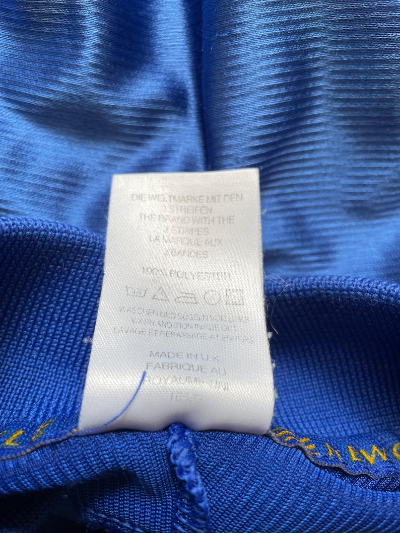 K&ouml;nnte beinhalten: Ein wei&szlig;es Etikett mit schwarzem Text auf blauem Hintergrund. Das Etikett lautet "Die Weltmarke mit den Streifen", "The Brand with the Stripes", "La Marque aux Bandes", "100% Polyester", "Waschen und Seselinordka", "Wash and Iron Inside Out", "Lavage et Repassage a l'envers", "Made in UK", "Fabrique au Royaume Uni".