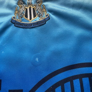 K&ouml;nnte beinhalten: Ein hellblaues Fu&szlig;balltrikot mit einem schwarz-wei&szlig;en Newcastle United Wappen auf der Vorderseite. Das Wappen zeigt zwei Seepferdchen und die Worte "Newcastle United".