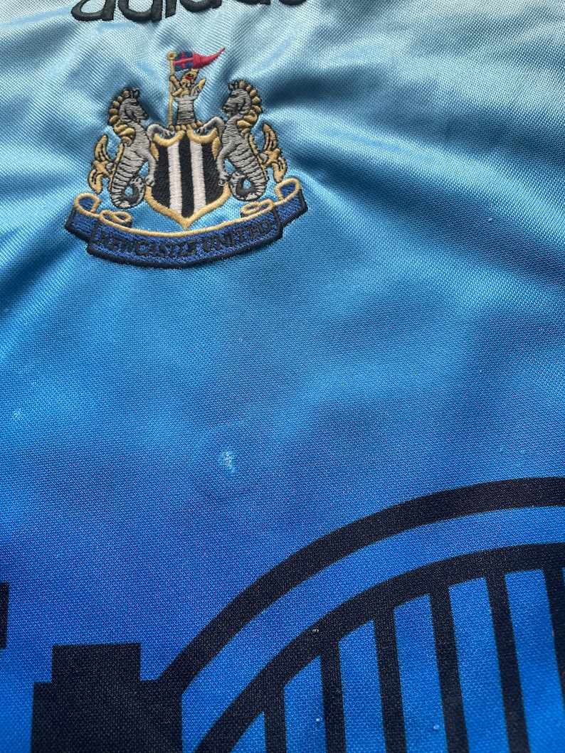 K&ouml;nnte beinhalten: Ein hellblaues Fu&szlig;balltrikot mit einem schwarz-wei&szlig;en Newcastle United Wappen auf der Vorderseite. Das Wappen zeigt zwei Seepferdchen und die Worte "Newcastle United".
