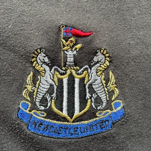 May include: Embroidered patch with the Newcastle United crest. The crest features a black and white shield with two gold seahorses on either side. The shield is topped with a gold crown and a red flag with a white cross. A blue ribbon with the words "Newcastle United" is wrapped around the bottom of the shield.