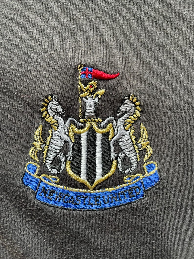 May include: Embroidered patch with the Newcastle United crest. The crest features a black and white shield with two gold seahorses on either side. The shield is topped with a gold crown and a red flag with a white cross. A blue ribbon with the words "Newcastle United" is wrapped around the bottom of the shield.