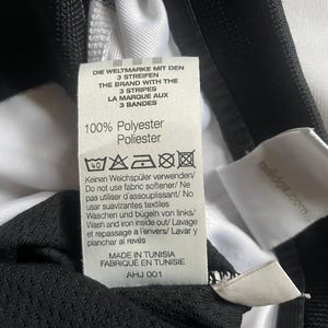 Puede incluir: Primer plano de una etiqueta de ropa con texto en varios idiomas, incluidos ingl&eacute;s, alem&aacute;n y franc&eacute;s. La etiqueta indica que la prenda es 100% poli&eacute;ster y proporciona instrucciones de cuidado. La etiqueta tambi&eacute;n dice "MADE IN TUNISIA".