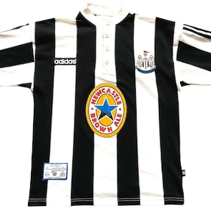May include: Black and white striped soccer jersey with the Adidas logo, Newcastle Brown Ale logo, and Newcastle United crest. The jersey has a button-up collar and short sleeves. A small tag is visible near the bottom.