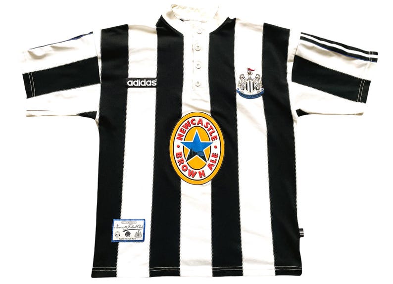 May include: Black and white striped soccer jersey with the Adidas logo, Newcastle Brown Ale logo, and Newcastle United crest. The jersey has a button-up collar and short sleeves. A small tag is visible near the bottom.