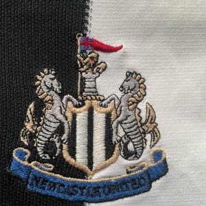 May include: Embroidered Newcastle United crest on a black and white striped background. The crest features two seahorses, a shield, and a banner with the text "NEWCASTLE UNITED" in blue.