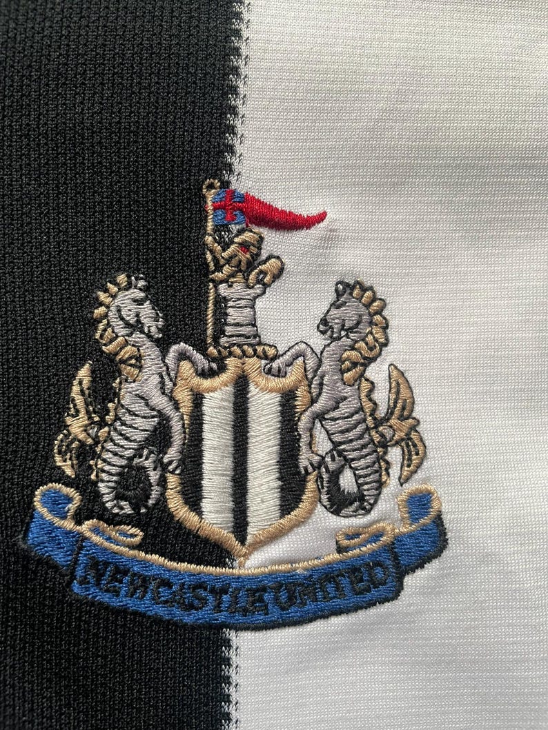 May include: Embroidered Newcastle United crest on a black and white striped background. The crest features two seahorses, a shield, and a banner with the text "NEWCASTLE UNITED" in blue.