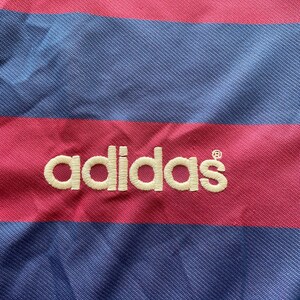 May include: Close-up of a striped fabric with alternating bands of pink and blue. The word "adidas" is embroidered in white across the fabric. The material appears to be a jersey or athletic wear.