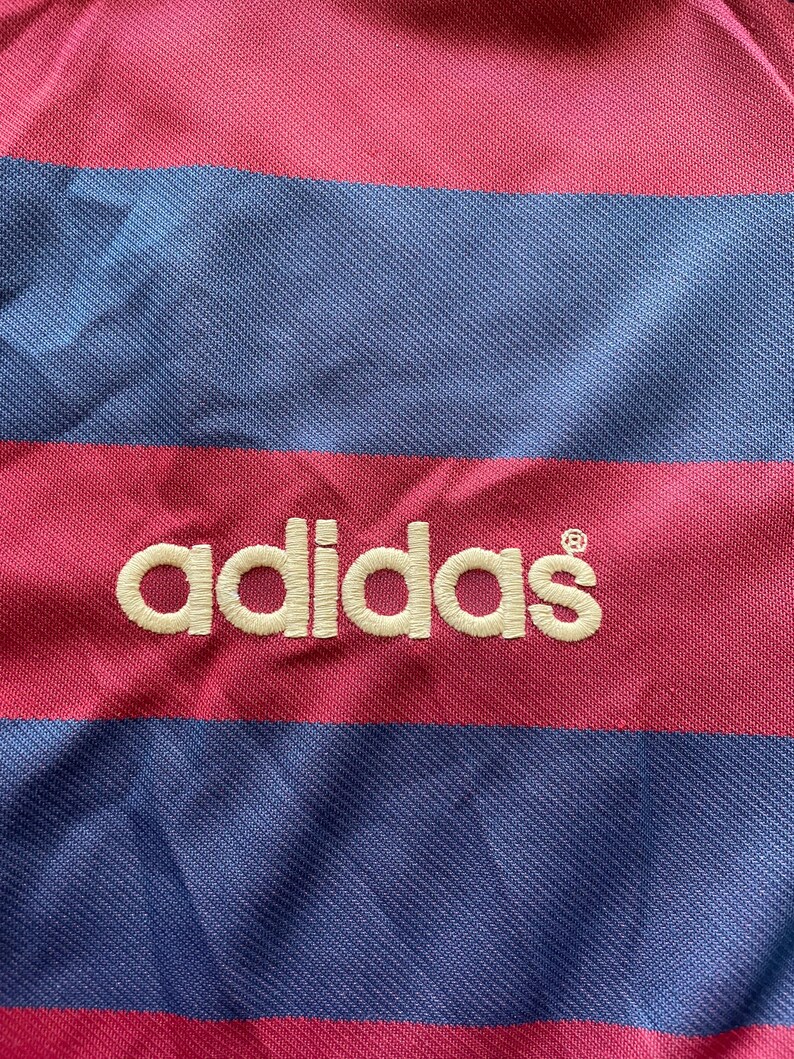 May include: Close-up of a striped fabric with alternating bands of pink and blue. The word "adidas" is embroidered in white across the fabric. The material appears to be a jersey or athletic wear.