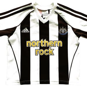 May include: White and black striped soccer jersey with brown accents and the text "northern rock". Features the Adidas logo and a team crest. The jersey has a V-neck collar.