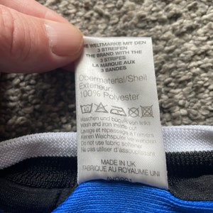 May include: A white tag with black text detailing the material composition and care instructions for a garment. The tag reads "Obermaterial/Shell Exterieur: 100% Polyester" and includes washing and ironing instructions in German, English, and French. The tag also states "Made in UK Fabrique au Royaume Uni AVG901".