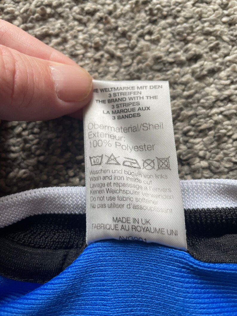 May include: A white tag with black text detailing the material composition and care instructions for a garment. The tag reads "Obermaterial/Shell Exterieur: 100% Polyester" and includes washing and ironing instructions in German, English, and French. The tag also states "Made in UK Fabrique au Royaume Uni AVG901".