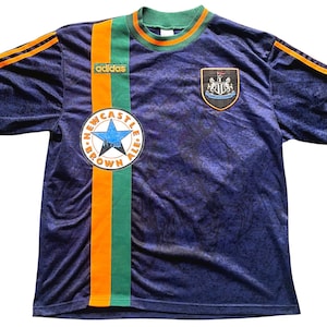 May include: Navy blue Adidas soccer jersey with orange and green accents. Features the Newcastle Brown Ale logo and a team crest. The jersey has orange stripes on the sleeves and a green collar.