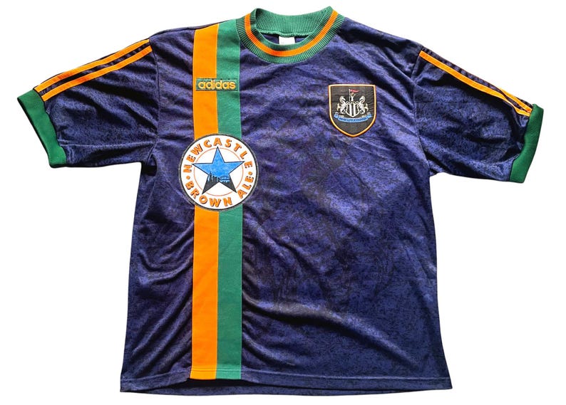 May include: Navy blue Adidas soccer jersey with orange and green accents. Features the Newcastle Brown Ale logo and a team crest. The jersey has orange stripes on the sleeves and a green collar.